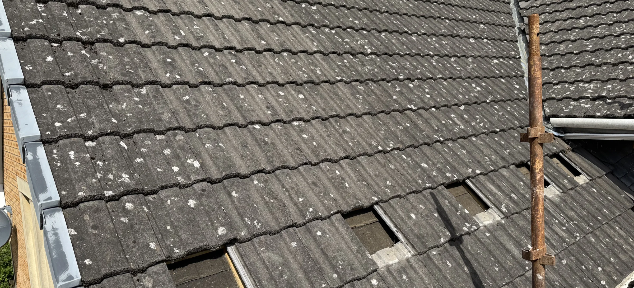 Several roof tiles lifted or removed in a row at mounting positions on a tiled pitched roof, exposing dark rectangular openings and scaffold access pole nearby; preparatory stage.
