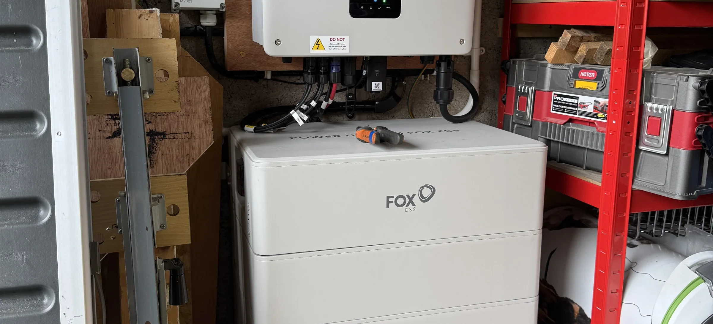 Fox ESS inverter mounted on a blockwork wall above a stacked Fox battery unit inside an outbuilding; rotary AC isolator and multiple cable glands/terminations are visible — (stage: