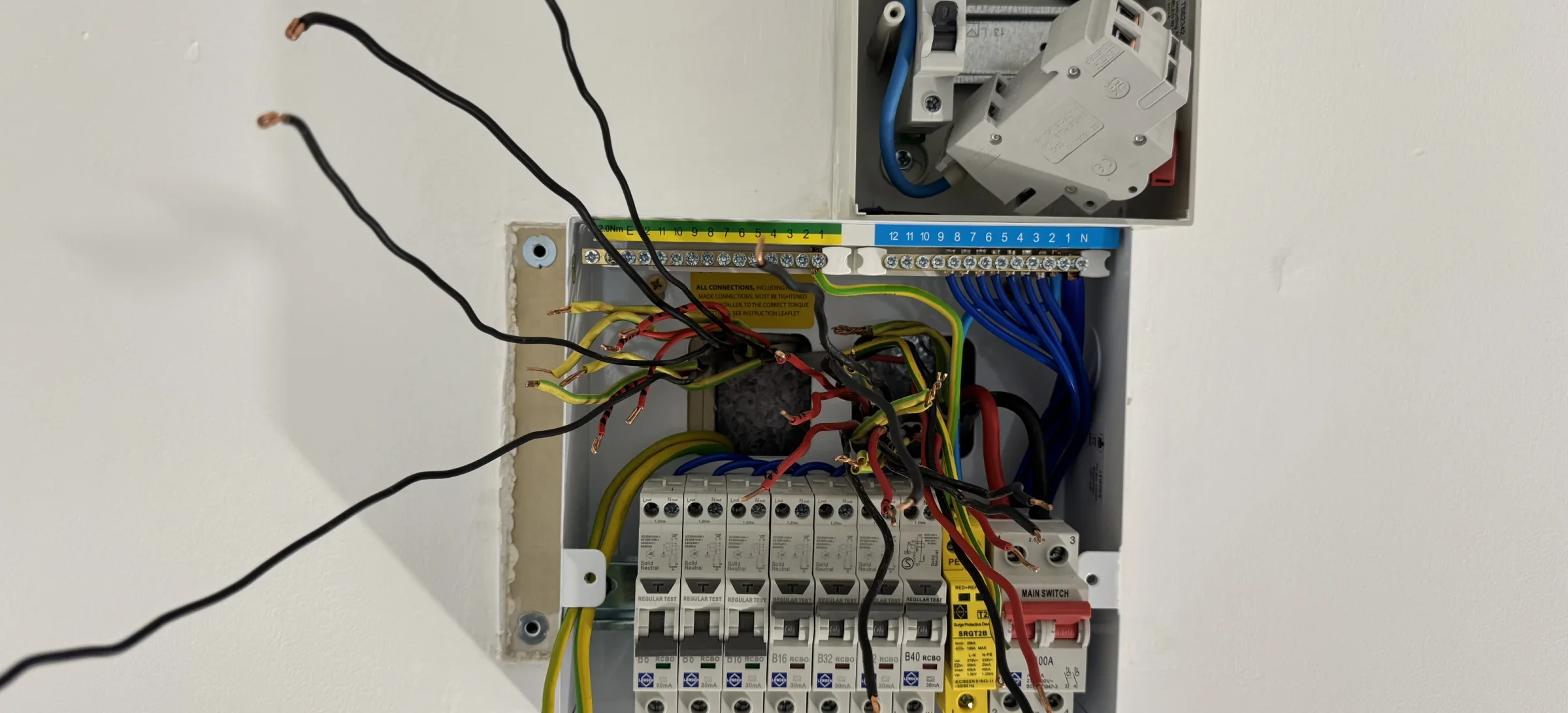 Wall cavity with the original consumer unit removed and multiple circuit conductors stripped and pulled forward for retermination; separate sub‑supply isolator and removed device