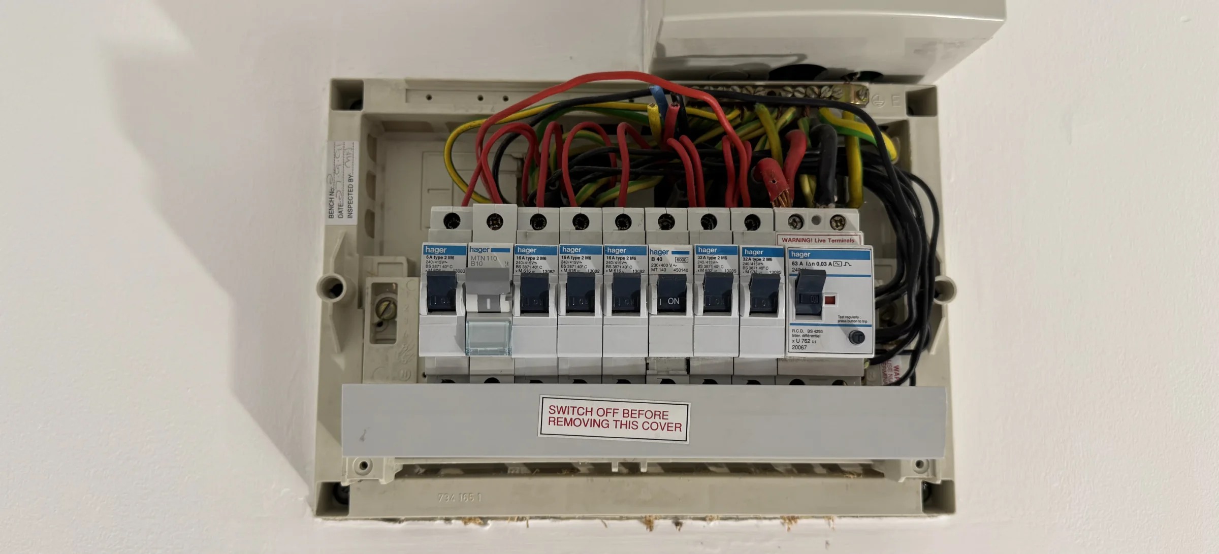 Open plastic consumer unit recessed in the wall beneath a separate sub‑supply isolator; original row of breakers and a residual current device visible with multiple live, neutral,和